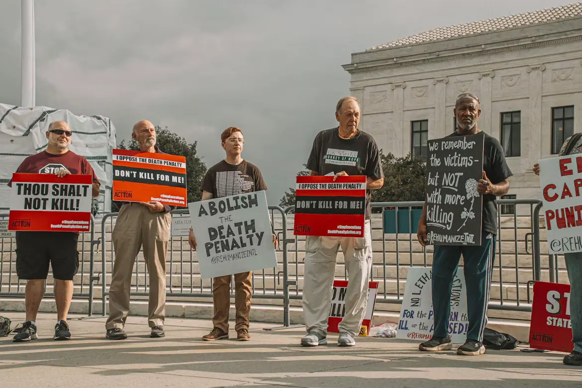 A group of people holding the signs of “Thou shalt not kill”, “Don’t kill for me”, “Abolish Death Penalty”, “We remember the victims - but no with more killing” in front of the fence.